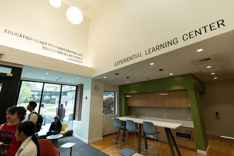Modern room labeled "Experiential Learning Center" with high ceiling, kitchen area, seating, and people gathered near large windows. Quote by John Dewey on wall.