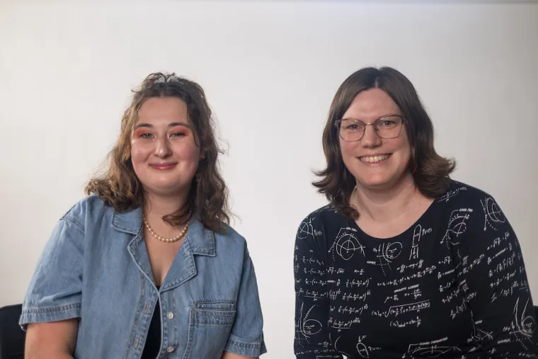 Two people are sitting and smiling at the camera. One wears a denim shirt and pearls; the other wears glasses and a shirt with scientific patterns.