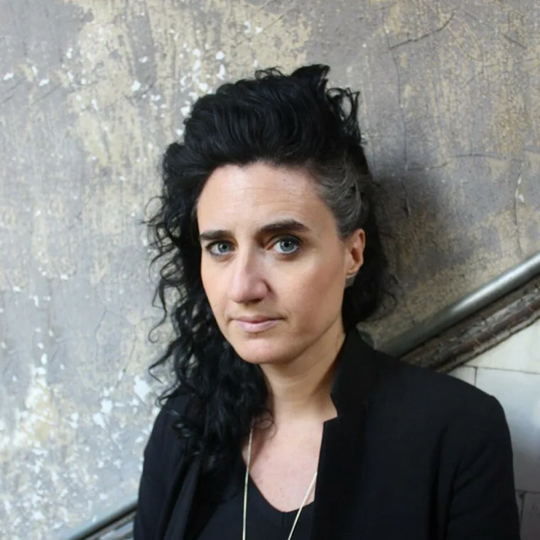Person with curly dark hair and a streak of gray stands against a textured wall, looking at the camera.