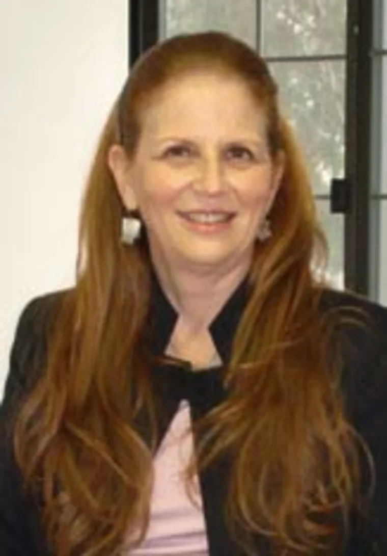Person with long hair smiling, wearing a black jacket and pink top, in front of a window.