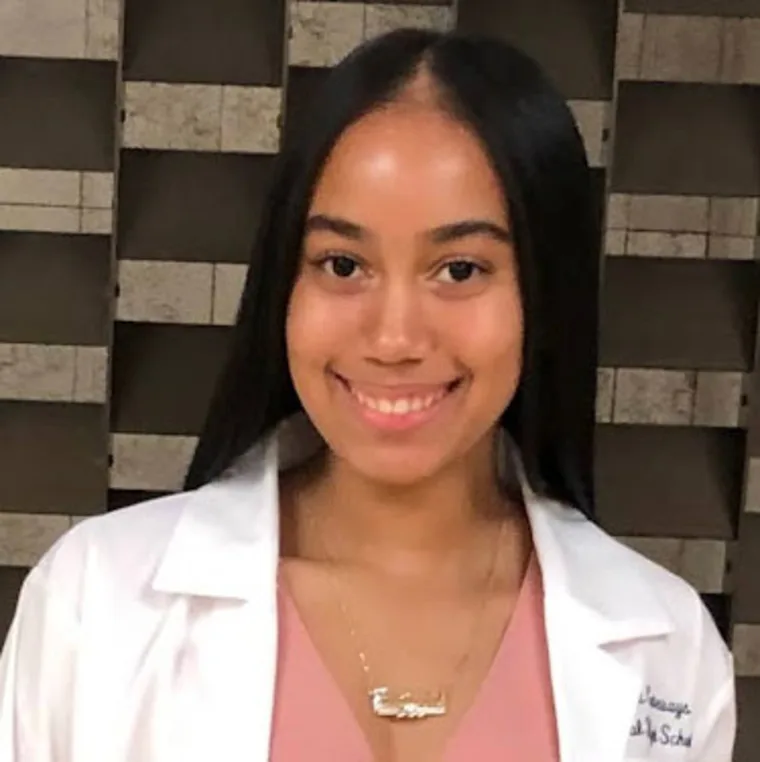 Person smiling, wearing a white coat and a name necklace. The background is a patterned stone wall.
