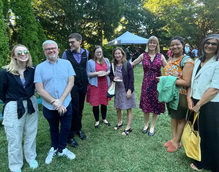Group of people smiling outdoors, standing on grass near a canopy. Trees and people in the background.