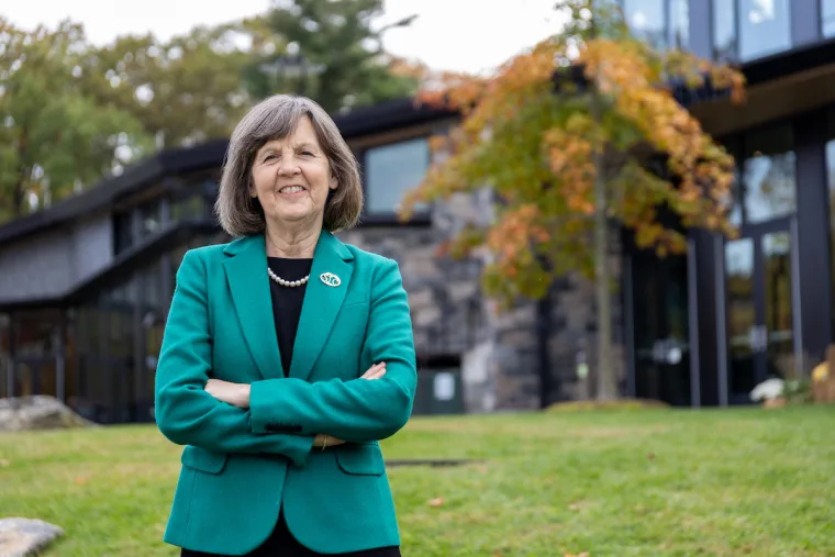 Person in a green blazer stands with arms crossed on grassy lawn, smiling. Modern building and autumn trees in the background.