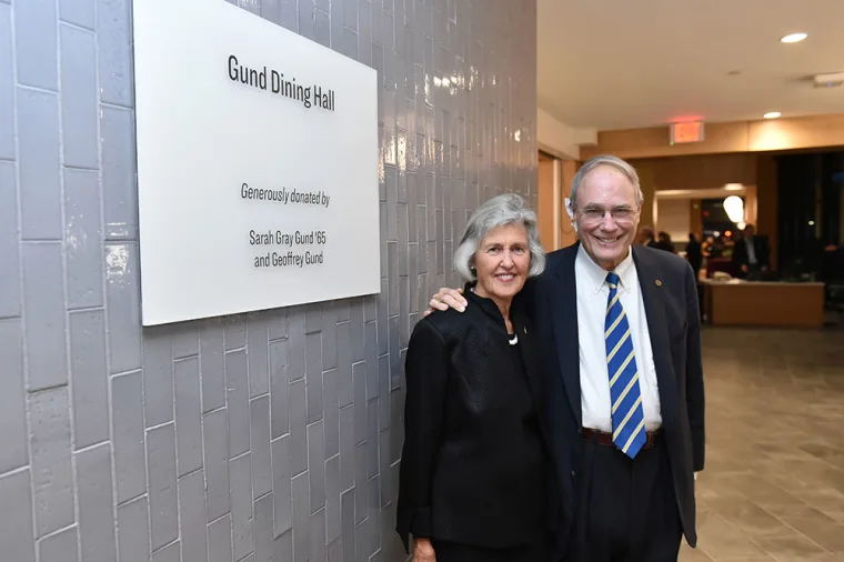 Two people stand smiling next to a sign reading "Gund Dining Hall." The sign mentions it was donated by Sarah Gray Gund and Geoffrey Gund.