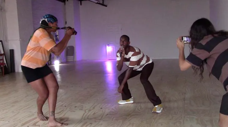 Three people in a studio. One operates a camera with a fabric strap, another strikes a pose, and the third takes a photo with a phone.