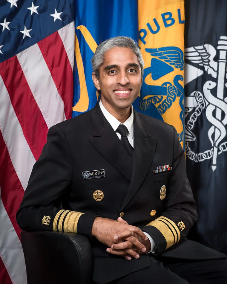 Officer in formal uniform with braided sleeves sits in front of flags, including the U.S. flag, smiling slightly, hands clasped.