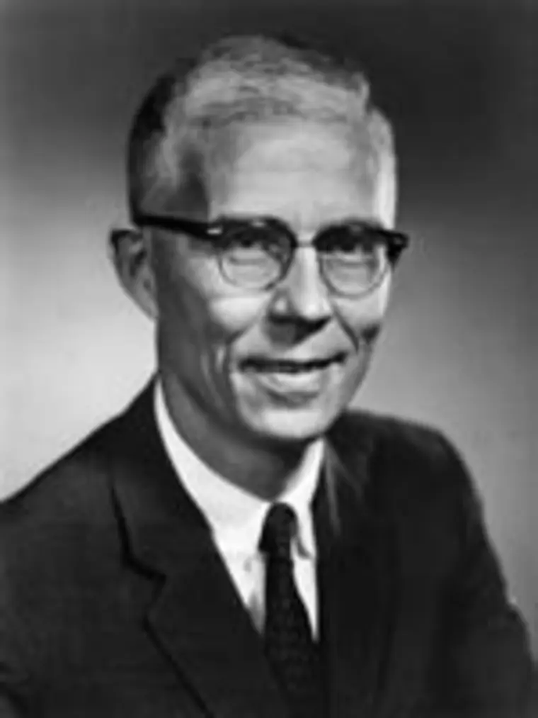 Person with short hair, wearing glasses and a suit, smiling slightly in a black and white portrait.