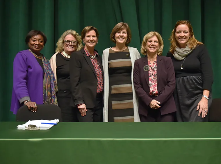 Six people stand together smiling in front of a green curtain, behind a table with a clipboard and pen.
