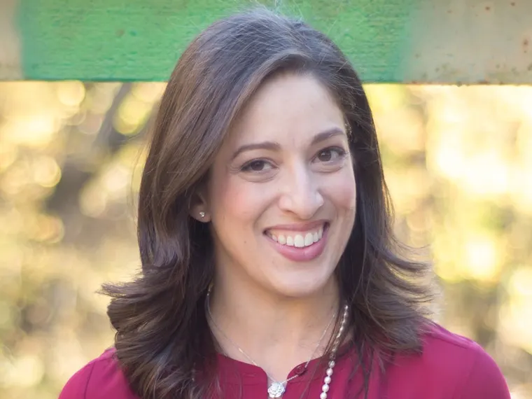 Person smiling outdoors with a blurred background of green and yellow hues, wearing a red top and necklace.