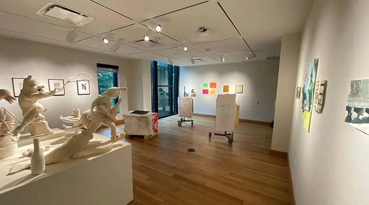 Art gallery with sculptures and paintings. Statues on left, colorful artworks on walls, wooden floor, track lighting, large windows in background.