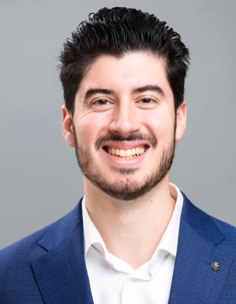 Person smiling, wearing a blue blazer and white shirt, with short dark hair and a beard, against a plain gray background.