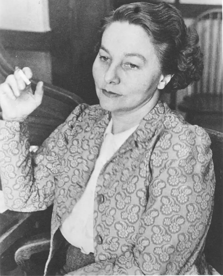 Person with short wavy hair in patterned jacket, sitting and holding a cigarette, appears in thought.