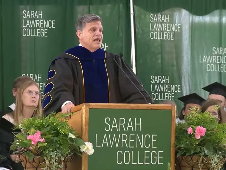 Person speaking at a podium with "Sarah Lawrence College" in the background. Audience members are visible in graduation attire.