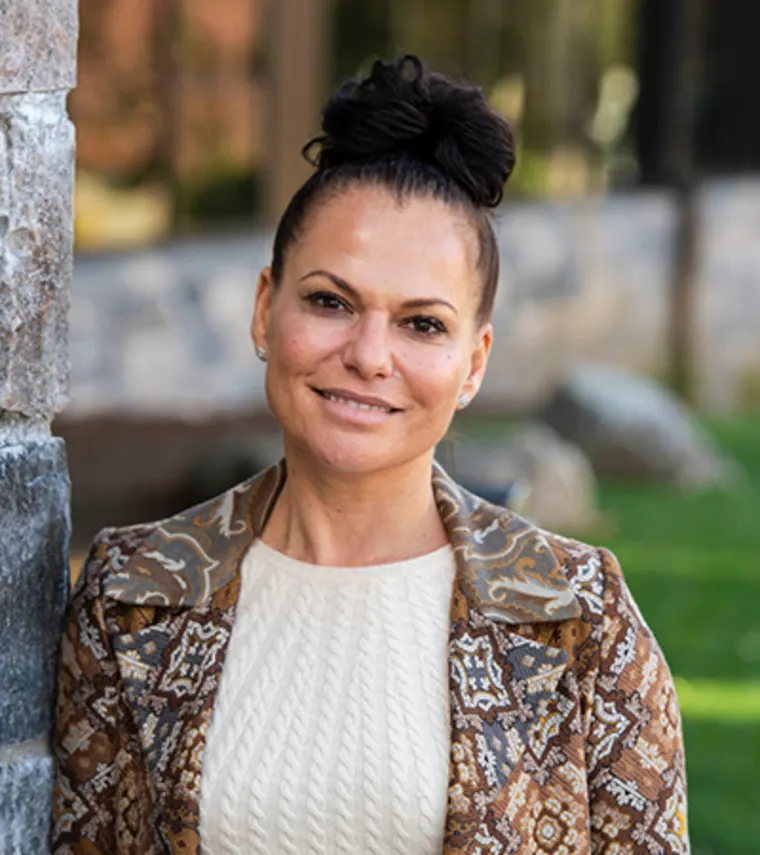 Person with hair in a bun, wearing a patterned jacket and light sweater, standing outdoors near a stone wall, grassy background.