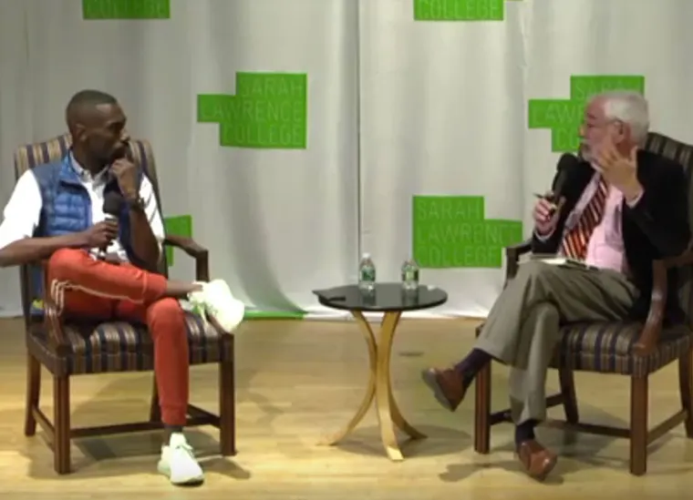 Two people sit in chairs on a stage with a "Sarah Lawrence College" backdrop, engaged in conversation. A small table with water bottles is between them.