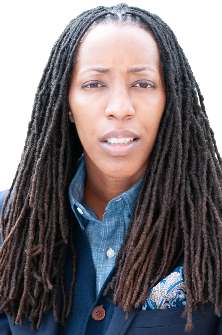 Person with long dreadlocks in a blue shirt and dark jacket, standing against a white background, looking forward with a neutral expression.