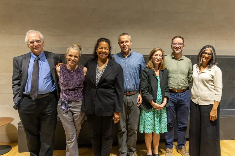 Seven people standing in a row, smiling, with a neutral wall and wooden floor as the background.
