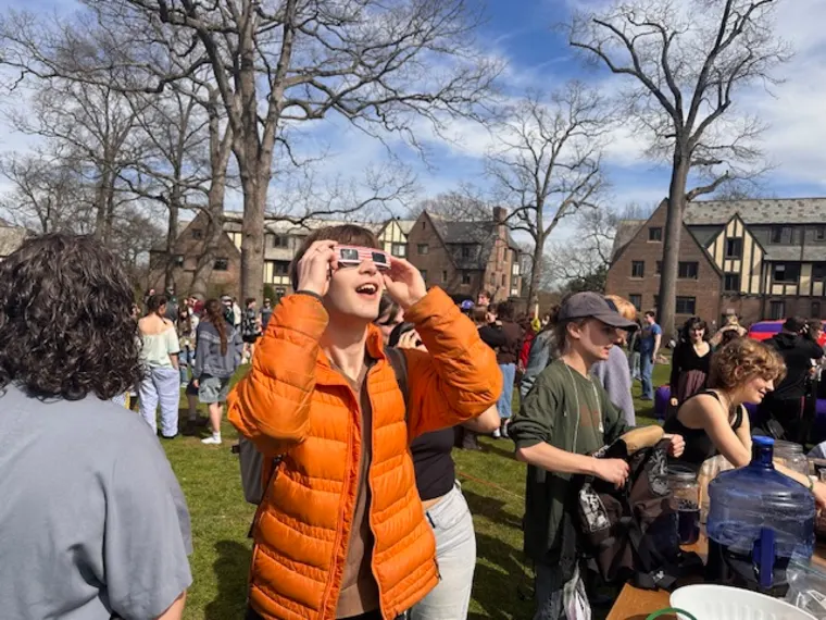 Person in orange jacket using glasses to view sky at an outdoor gathering, surrounded by people and buildings in the background.