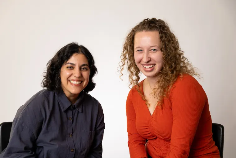 Two people seated and smiling at the camera, one with short dark hair in a dark shirt, the other with curly hair in an orange top.