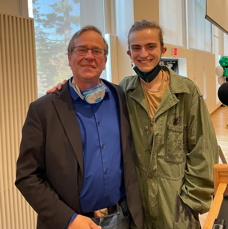 Two people smiling, one wearing a suit with an air mask, the other in a green jacket. They are indoors with large windows in the background.