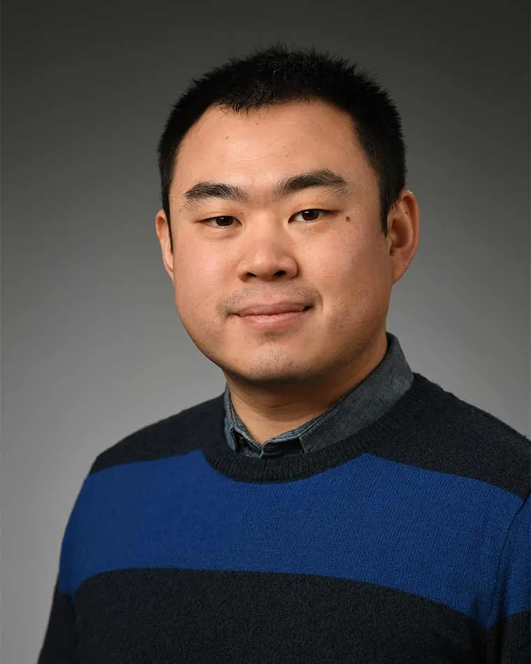 Person in a blue and black striped sweater, smiling slightly, with a gray background.