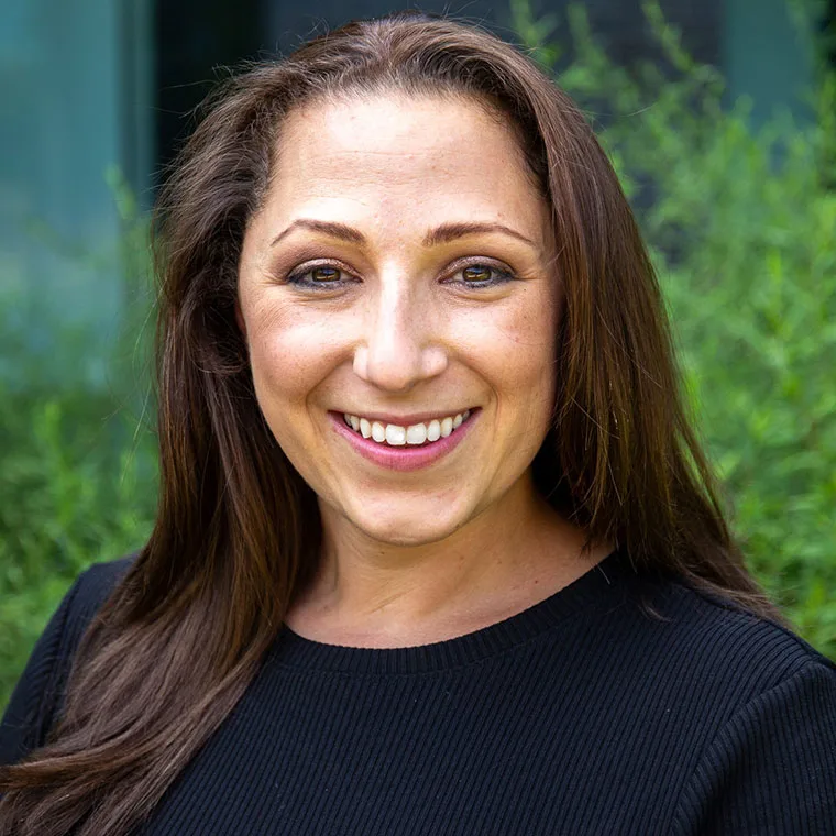 Person with long brown hair and a black top smiling in front of green foliage.