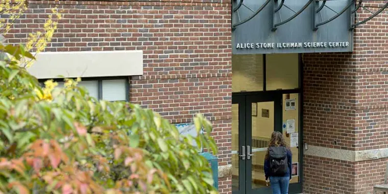 Person with a backpack entering Alice Stone Ilchman Science Center, surrounded by brick walls and foliage.