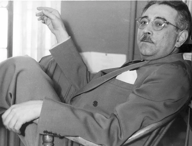 Person in a suit sits relaxed in a chair, looking towards the camera, with one arm raised and glasses on.