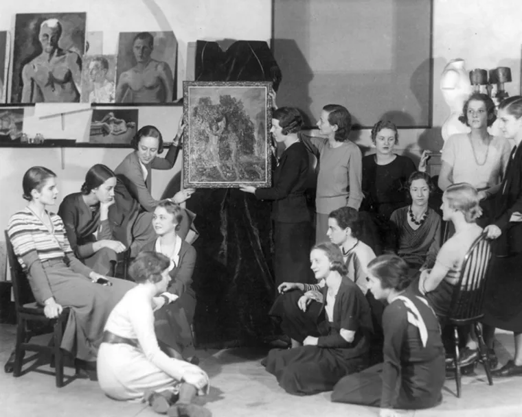A group of people gather in an art studio, inspecting a framed painting. Various paintings and sculptures surround the scene.
