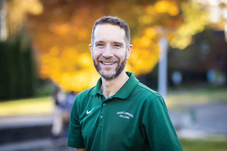 Person in a green "Sarah Lawrence College" polo shirt smiling, standing outdoors with vibrant autumn foliage in the background.