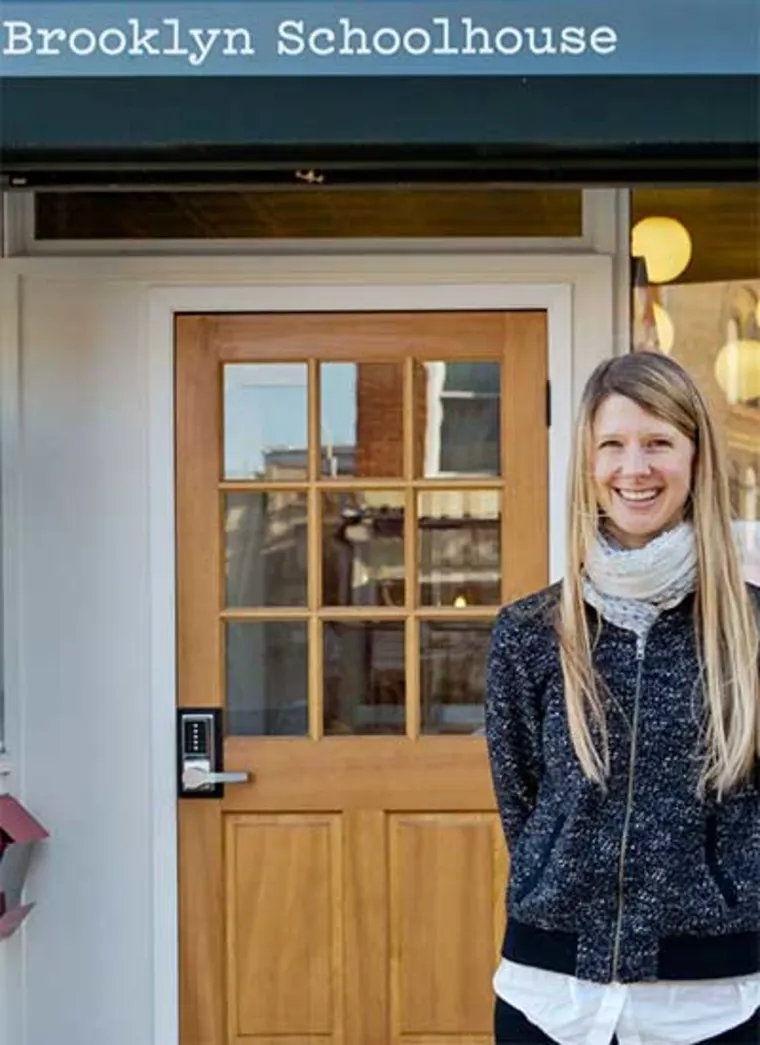 Person smiling in front of a wooden door with a sign above reading "Brooklyn Schoolhouse."