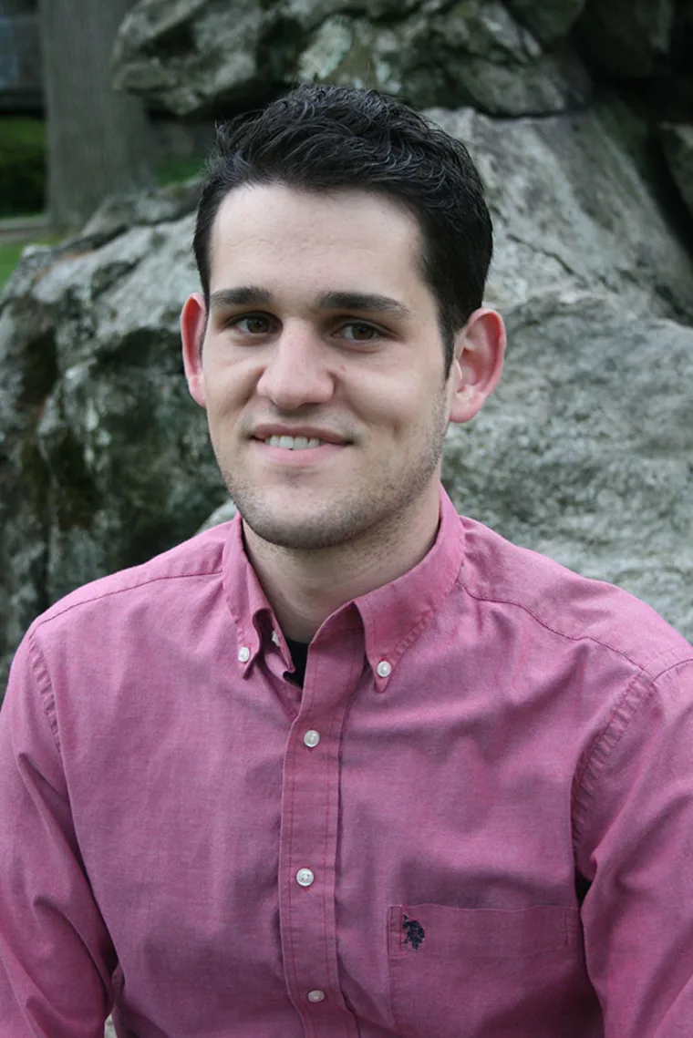 Person in a pink button-up shirt smiling, standing outdoors in front of large rocks.