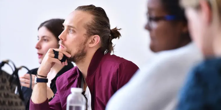 People seated and focused in a classroom setting, one in the foreground with a ponytail and burgundy jacket, chin resting on hand.