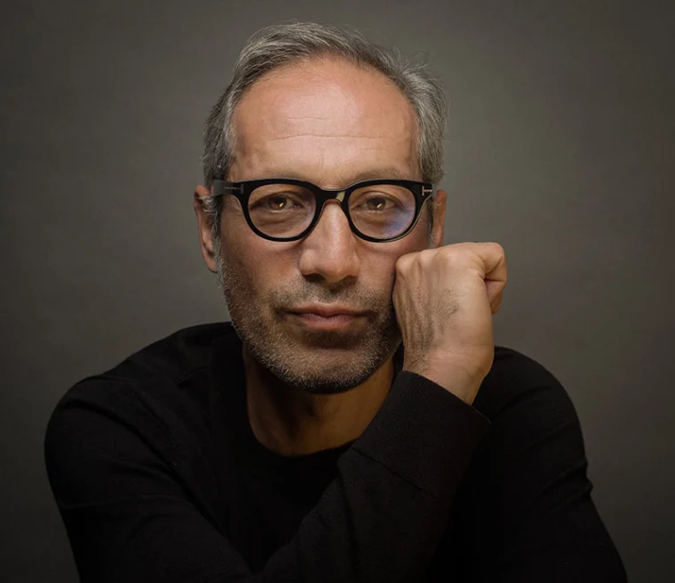 Person with short hair wearing glasses, resting chin on hand, in a dark shirt against a gray background.
