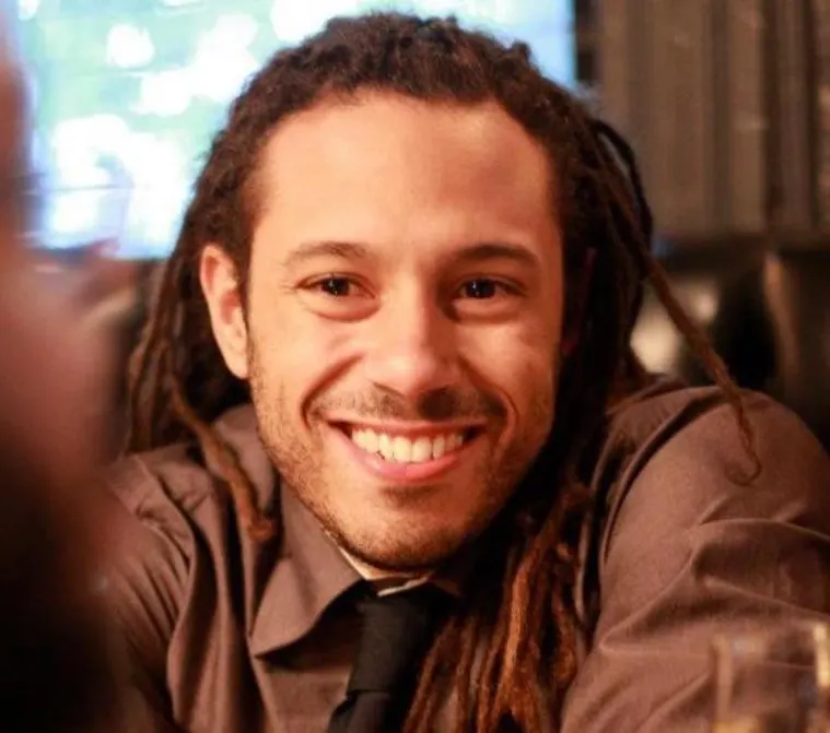 Person with long hair and a beard smiling, wearing a shirt and tie, sitting indoors with a blurred background.