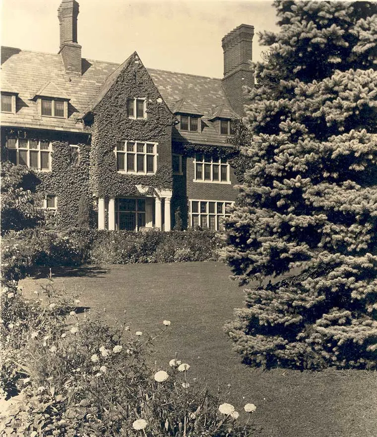 Large ivy-covered mansion with multiple chimneys and windows, surrounded by manicured garden, flowers, and a tall evergreen tree.