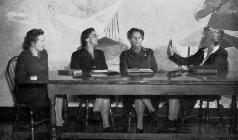 Four people sit at a table with books, conversing. The background features abstract wall art.