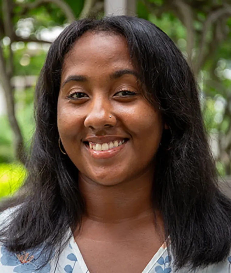 Person smiling outdoors with long dark hair, wearing a light blue and white patterned top, and a small nose ring. Greenery in the background.