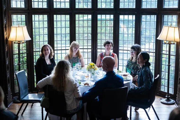 Seven people sitting around a dining table with a white tablecloth and flower centerpiece, in a room with large windows and two floor lamps.