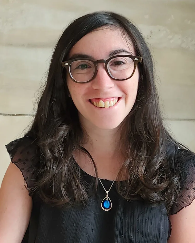 Person with long hair and glasses smiles, wearing a black shirt with a blue pendant necklace.