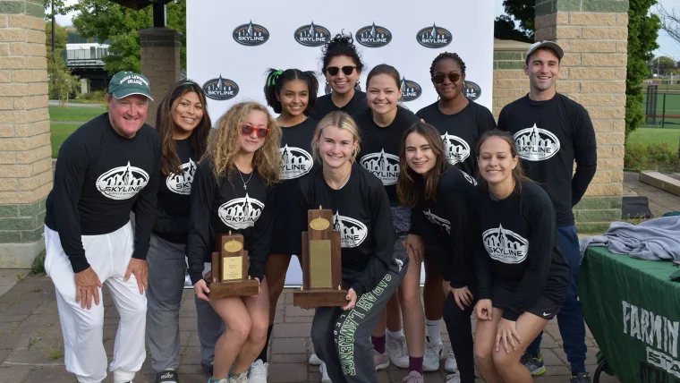 Group of people in matching shirts smiling at the camera, two are holding trophies. A branded backdrop and a table are in the background.