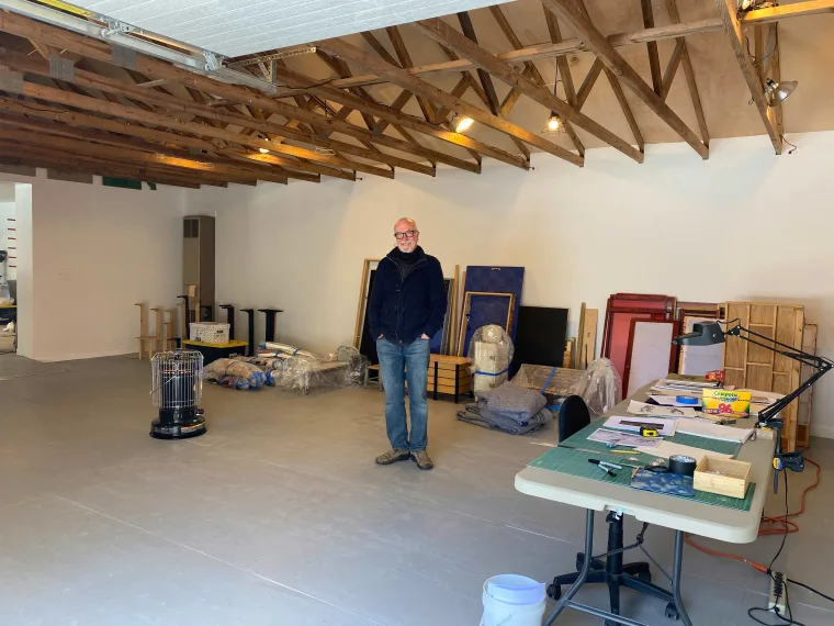 Person standing in a large room with exposed wooden beams, surrounded by art supplies, tables, a chair, and unpacked materials.