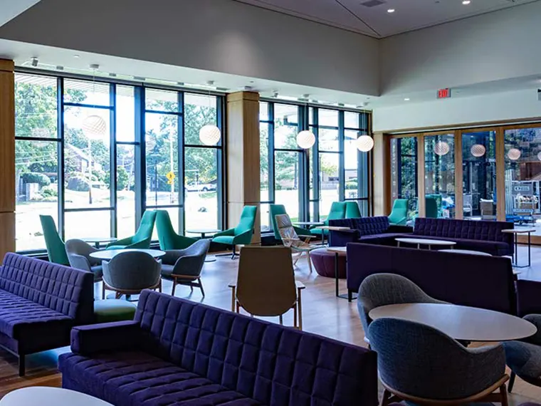Modern lounge area with purple sofas, green chairs, round tables, and large windows providing natural light.