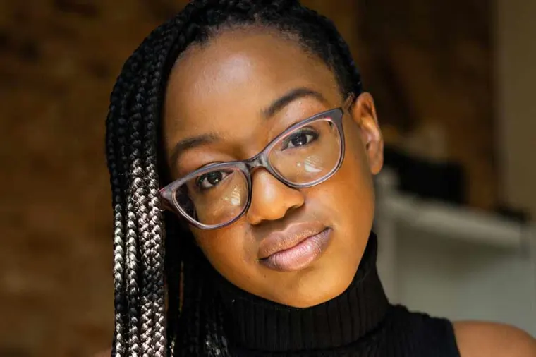 A person with braided hair and glasses looks at the camera, wearing a black sleeveless turtleneck against a blurred background.