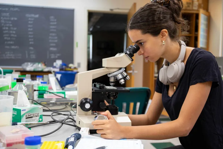 Person using a microscope in a laboratory with various supplies and a chalkboard in the background.