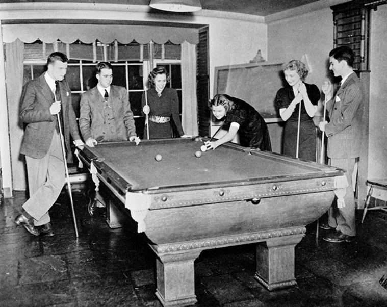 Six people gather around a pool table, one person is taking a shot while the others watch and hold cues.