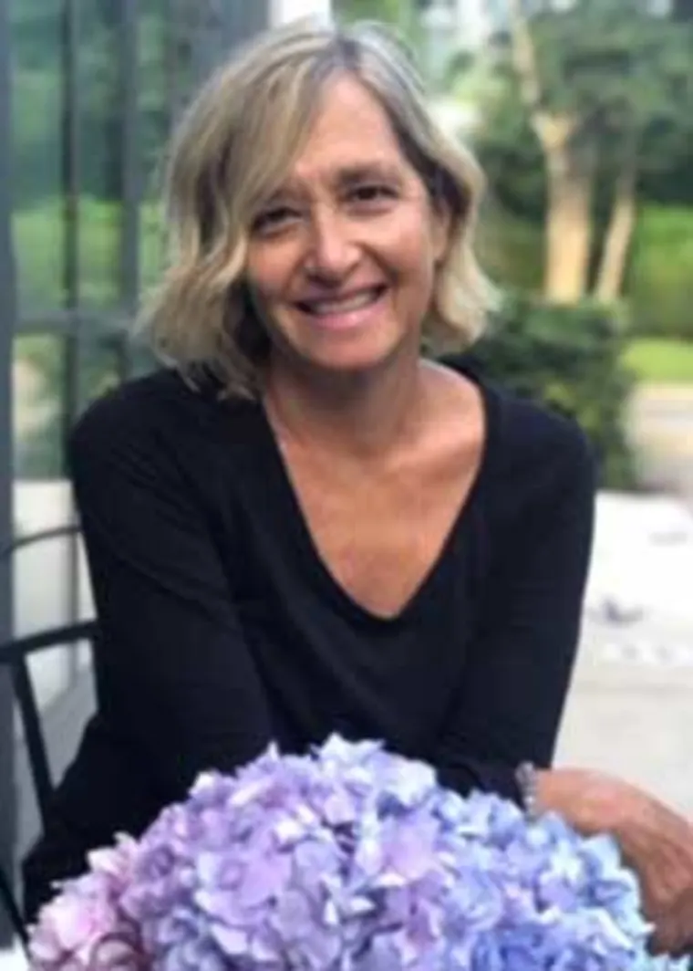 Person with short, wavy hair, smiling, wearing a black top, seated beside a bouquet of purple flowers outdoors.