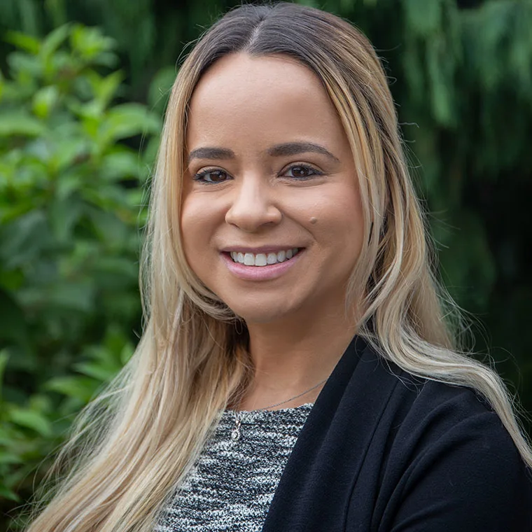 Person smiling outdoors with long hair, wearing a black jacket and gray top. Green foliage in the blurred background.