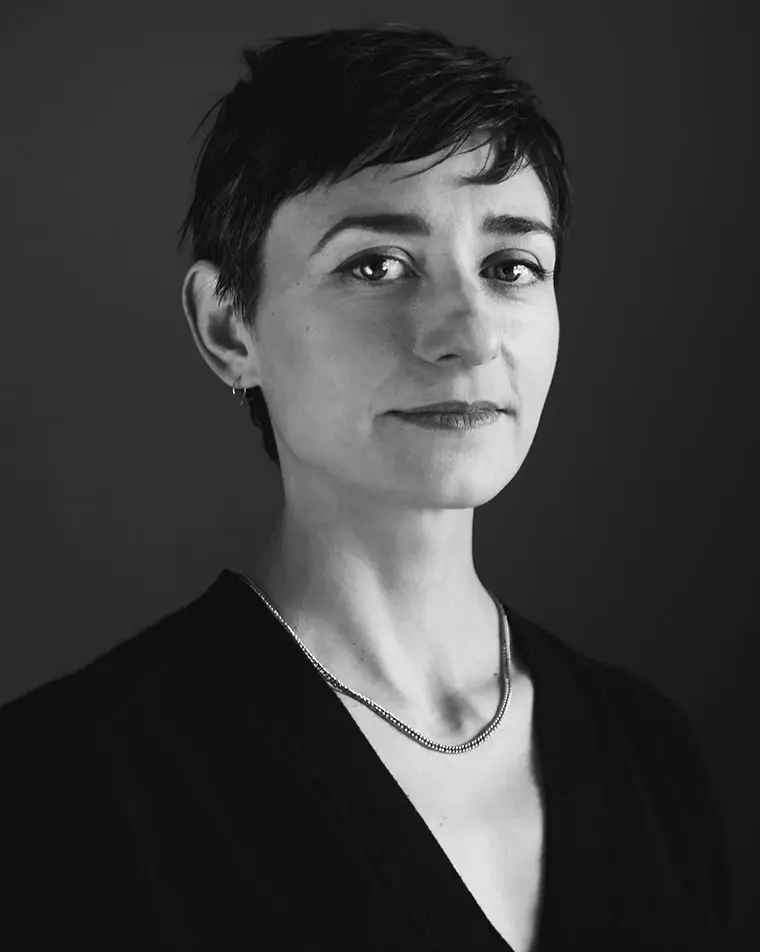 Person with short hair and necklace, gazing at the camera in a black and white portrait.