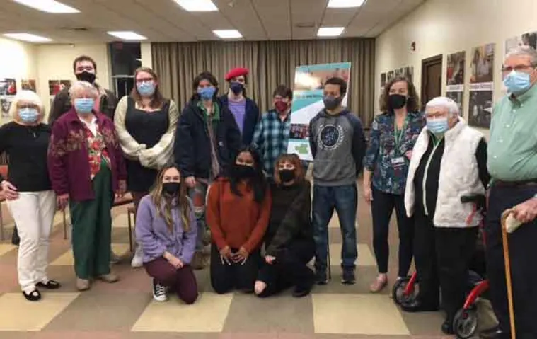 Group of people wearing masks pose indoors in front of a poster, with some seated and others standing, in a well-lit room.
